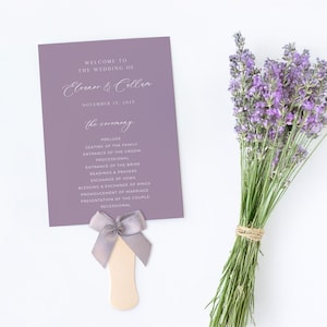 May include: Purple wedding ceremony program fan with a gray satin bow. The program lists the order of events for the wedding ceremony, including the prelude, seating of the family, entrance of the groom, processional, entrance of the bride, readings and prayers, exchange of vows, blessing and exchange of rings, pronouncement of marriage, presentation of the couple, and recessional. The text "Welcome to the wedding of Eleanor & Cellen, November 15, 2025" is at the top of the program.
