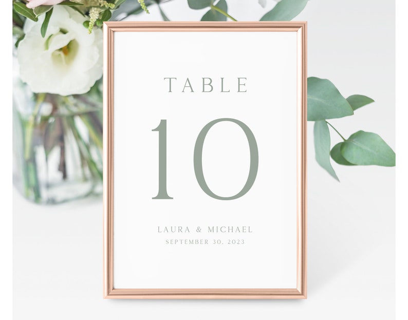 May include: A rose gold table number card with the number 10 in a light green color. The text "TABLE" is at the top of the card and the text "LAURA & MICHAEL SEPTEMBER 30, 2023" is at the bottom.