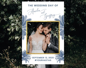 May include: A white wedding photo booth sign with a gold frame and blue palm tree illustrations. The sign reads "The Wedding Day of Amelia and Geoffrey" and "September 12, 2020 #YOURHASTAG".