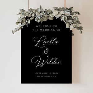 May include: Black wedding sign with white text that reads "Welcome to the wedding of Luella & Wilder, September 15, 2024, San Francisco, CA". The sign is decorated with eucalyptus greenery.