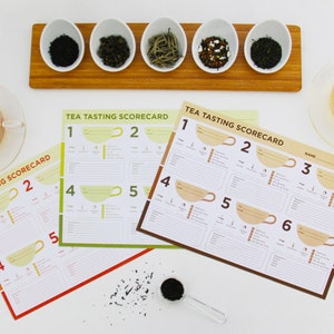 Könnte beinhalten: Draufsicht auf ein Tee-Tasting-Setup. Enthält kleine weiße Schalen mit verschiedenen losen Teeblättern, Tee-Tasting-Scorecards und klare Teetassen aus Glas, gefüllt mit aufgebrühtem Tee. Die Scorecards tragen den Text "TEA TASTING SCORECARD".