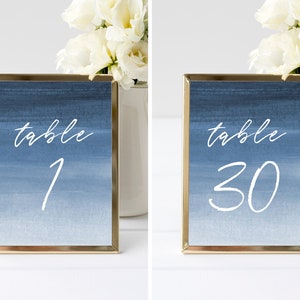 May include: Two gold-framed table number signs with a blue watercolor background. The numbers are white and handwritten. The first sign says "table 1" and the second sign says "table 30".