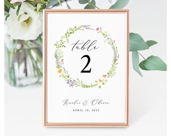 Pode incluir: Um cartão de número de mesa branco com um design de coroa de flores. O cartão diz "Mesa 2" e inclui os nomes "Rosalie & Oliver" e a data "10 de abril de 2022".