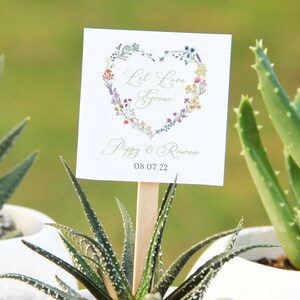 May include: A white card with a floral heart design and the text "Let Love Grow" in green script. Below the heart is the text "Poppy & Rowan" and the date "08.07.22".