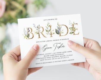 May include: A white bridal shower invitation with gold lettering that reads "Let's Shower the Bride". The invitation features a floral design with green leaves and white flowers. The text also includes the bride's name, the date, time, and location of the shower, as well as RSVP information.
