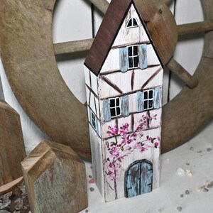 Könnte beinhalten: Ein handbemaltes Holzhaus mit braunem Dach, weißen Wänden und blauen Fensterläden. Das Haus hat ein Blumendesign und eine blaue Tür. Es ist ein dekorativer Gegenstand.