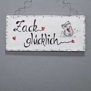 Könnte beinhalten: Ein weißes Holzschild mit schwarzer Schrift, auf dem "Zack glücklich" steht, mit einer kleinen grauen Katze, die ein rotes Herz hält. Das Schild ist mit kleinen roten Herzen verziert.