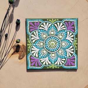 May include: A green square canvas with a mandala design painted in white, blue, green, and purple dots.