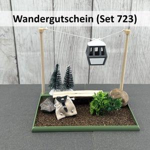 Könnte beinhalten: Ein Miniatur-Diorama mit einer schwarz-weißen Seilbahn, die zwischen zwei Holzpfählen hängt. Die Szene umfasst Miniaturbäume, eine kleine Bank, einen Stein und eine kleine Tasche. Der Text "Wandergutschein (Set 723)" wird oben angezeigt.