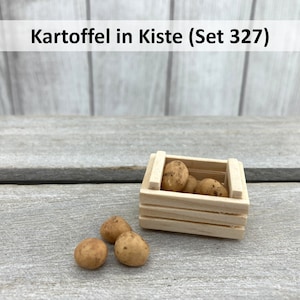 Könnte beinhalten: Eine kleine Holzkiste gefüllt mit braunen Kartoffeln. Drei weitere Kartoffeln liegen auf einer Holzoberfläche neben der Kiste. Die Kiste trägt den Aufdruck "Kartoffel in Kiste (Set 327)".