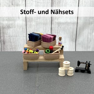 Könnte beinhalten: Eine Miniatur-Ausstellung von Nähzubehör, darunter Stoffbündel in Weidenkörben, Garnrollen und eine kleine Nähmaschine. Der Text "Stoff- und Nähsets" wird oben angezeigt.