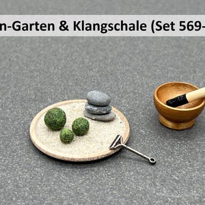 Könnte beinhalten: Ein Miniatur-Zen-Garten-Set mit einer Holzschale, einem kleinen Rechen und drei grünen Moosbällen. Das Set enthält außerdem einen Stapel grauer Steine und einen Holzstab zum Anschlagen der Schale. Das Set ist mit "Zen-Garten & Klangschale (Set 569-A)" beschriftet.