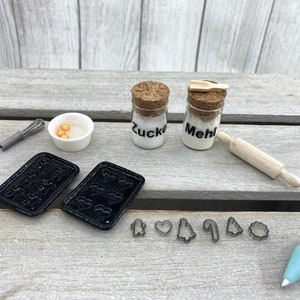 K&ouml;nnte beinhalten: Miniatur-Backset mit schwarzen Backblechen, Ausstechern, einem Schneebesen, einem Nudelholz und Gl&auml;sern mit der Aufschrift "Zucker" und "Mehl". Eine kleine Sch&uuml;ssel enth&auml;lt gelbe Kugeln. Ein blauer Spritzbeutel ist ebenfalls enthalten.