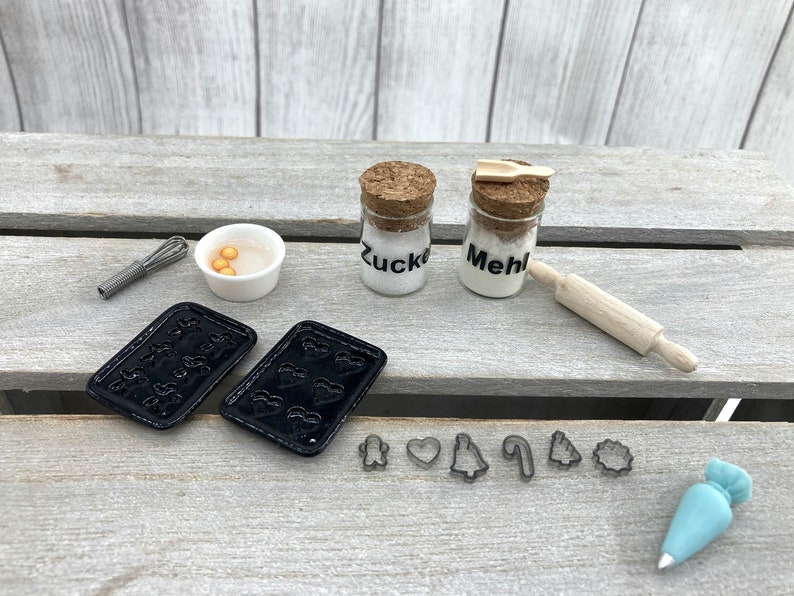 K&ouml;nnte beinhalten: Miniatur-Backset mit schwarzen Backblechen, Ausstechern, einem Schneebesen, einem Nudelholz und Gl&auml;sern mit der Aufschrift "Zucker" und "Mehl". Eine kleine Sch&uuml;ssel enth&auml;lt gelbe Kugeln. Ein blauer Spritzbeutel ist ebenfalls enthalten.