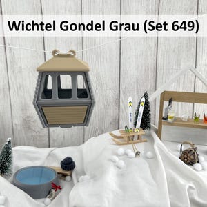 Könnte beinhalten: Eine Miniatur-Skigebietszene mit einer grauen und beigen Gondel, die an einer Schnur hängt. Die Szene umfasst eine kleine Bar, Skier, einen Whirlpool und ein schwarzes Fass. Der Text "Wichtel Gondel Grau (Set 649)" steht oben.