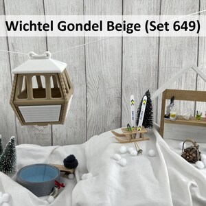 Könnte beinhalten: Eine beige und weiße Miniatur-Skiliftkabine hängt über einer Winterszene mit einer kleinen Bar, Skiern und einem Whirlpool. Die Kabine hat ein weißes Dach und einen beigen Körper. Die Szene umfasst Miniaturbäume und Schneebälle.