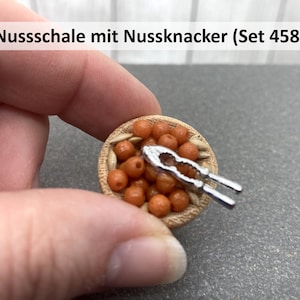 Könnte beinhalten: Eine Miniatur-Holzschale gefüllt mit orangen Nüssen und einem silbernen Nussknacker. Die Schale ist mit "Nussschale mit Nussknacker (Set 458)" beschriftet.