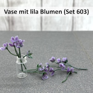Könnte beinhalten: Eine kleine Glasvase mit lila Blumen. Die Vase enthält mehrere Stiele lavendelfarbener Blumen mit grünen Stielen und Blättern. Zusätzliche Zweige derselben Blumen sind in der Nähe verstreut. Der Text "Vase mit lila Blumen (Set 603)" befindet sich oben.