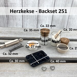 May include: A miniature baking set with a rolling pin, a whisk, a bowl of yellow dough, a baking sheet, a sifter, and two jars labelled "Zucker" and "Mehl". The set is labelled "Herzkekse - Backset 251".