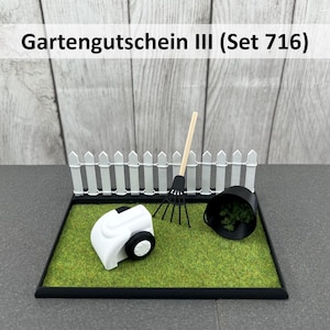 Könnte beinhalten: Eine Miniaturgartenszene mit einem weißen Rasenmäher, einer schwarzen Harke und einem schwarzen Eimer mit grünem Laub. Ein weißer Zaun grenzt an den grünen Rasen. Der Text "Gartengutschein III (Set 716)" wird oben angezeigt.