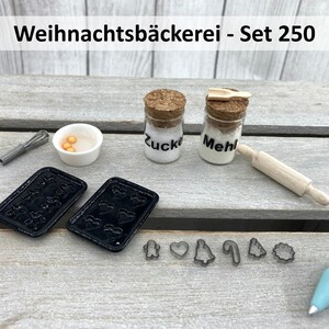 K&ouml;nnte beinhalten: Miniatur-Backset mit dem Text "Weihnachtsb&auml;ckerei - Set 250". Enth&auml;lt zwei schwarze Backbleche, kleine Gl&auml;ser mit der Aufschrift "Zucker" und "Mehl", ein Nudelholz, Ausstecher, einen Schneebesen, eine Sch&uuml;ssel mit gelben Kugeln und einen Spritzbeutel.