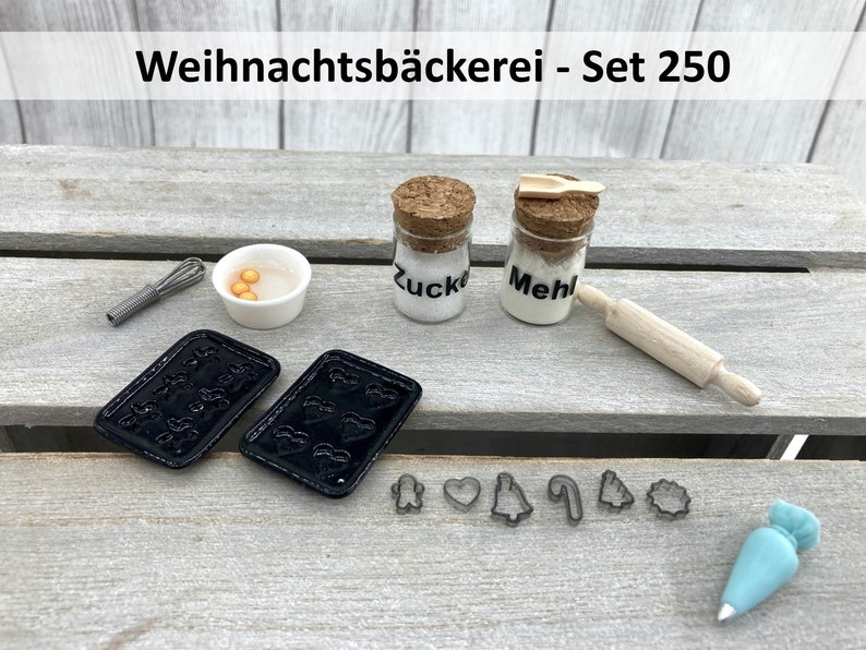 K&ouml;nnte beinhalten: Miniatur-Backset mit dem Text "Weihnachtsb&auml;ckerei - Set 250". Enth&auml;lt zwei schwarze Backbleche, kleine Gl&auml;ser mit der Aufschrift "Zucker" und "Mehl", ein Nudelholz, Ausstecher, einen Schneebesen, eine Sch&uuml;ssel mit gelben Kugeln und einen Spritzbeutel.