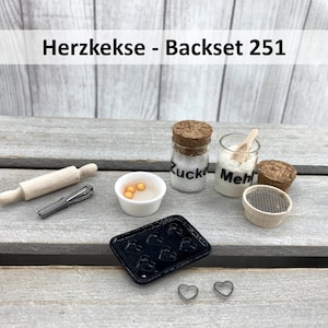 May include: A miniature baking set with the text "Herzkekse - Backset 251" on a wooden surface. The set includes a rolling pin, a whisk, a small bowl with yellow egg-shaped beads, two glass jars labelled "Zucker" and "Mehl", a small wooden sifter, and a black baking tray with heart-shaped moulds. There are also two small metal heart-shaped cookie cutters.