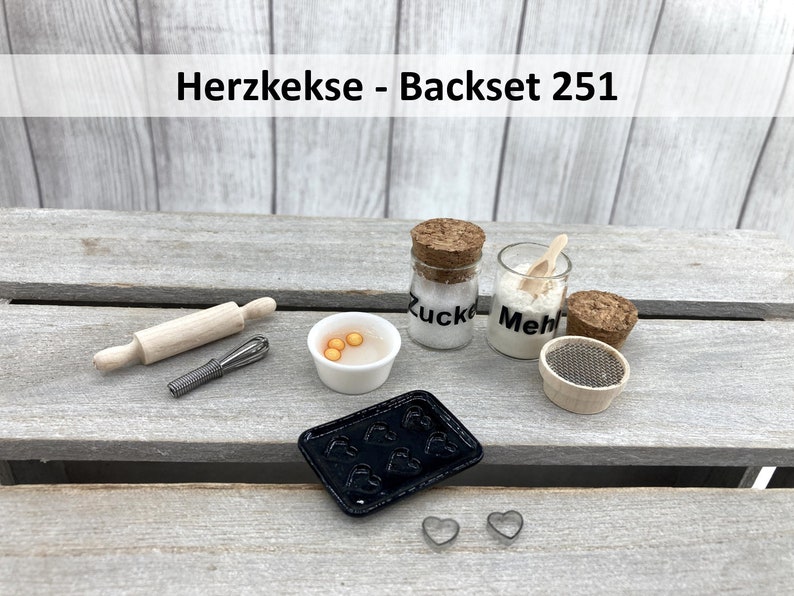 May include: A miniature baking set with the text "Herzkekse - Backset 251" on a wooden surface. The set includes a rolling pin, a whisk, a small bowl with yellow egg-shaped beads, two glass jars labelled "Zucker" and "Mehl", a small wooden sifter, and a black baking tray with heart-shaped moulds. There are also two small metal heart-shaped cookie cutters.