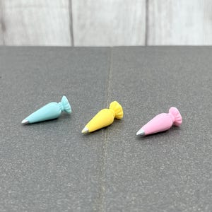 K&ouml;nnte beinhalten: Drei Miniatur-Spritzbeutel in Pastellfarben: Hellblau, Gelb und Rosa. Jeder Beutel hat eine silberne Spitze und einen ger&uuml;schten oberen Rand. Die Beutel sind auf einer dunkelgrauen Oberfl&auml;che angeordnet.