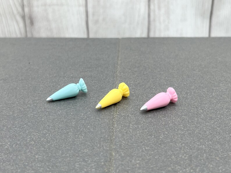 K&ouml;nnte beinhalten: Drei Miniatur-Spritzbeutel in Pastellfarben: Hellblau, Gelb und Rosa. Jeder Beutel hat eine silberne Spitze und einen ger&uuml;schten oberen Rand. Die Beutel sind auf einer dunkelgrauen Oberfl&auml;che angeordnet.
