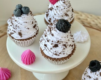 Peut inclure: Plusieurs cupcakes avec glaçage blanc, copeaux de chocolat et garnitures de fruits, dont des myrtilles, des framboises et des mûres. Les cupcakes sont présentés sur un présentoir à gâteaux blanc et une surface brun clair.
