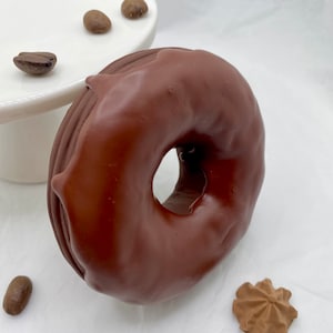 Pode incluir: Um donut coberto de chocolate com um buraco no centro, rodeado por grãos de café e um pequeno biscoito. O donut é de cor castanho escuro, e os grãos de café são de um tom castanho mais claro. O biscoito é de cor castanho claro.