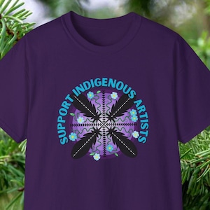 Pode incluir: Camiseta roxa com um design gráfico de um círculo com o texto "Support Indigenous Artists" em fonte azul. O círculo apresenta um design estilizado de penas e flores.