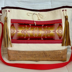 May include: Cream tote bag with a red adjustable strap. The bag features a geometric patterned panel in shades of red, yellow, and pink. It has a cork base and tan tassels. The bag's trim is a deep burgundy.