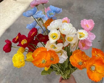 May include: A bouquet of colorful artificial poppy flowers in shades of pink, blue, red, orange, yellow, and white. The flowers have green stems and are held together. The petals have a delicate, textured appearance.