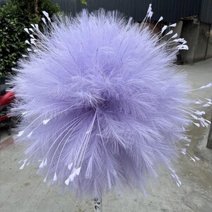 May include: A large, round decorative piece composed of soft, lavender-colored feathers. Delicate white accents extend outward from the main body, creating a unique, airy appearance. The item is likely used for floral arrangements or event decor.