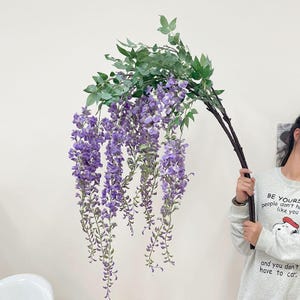 Op de afbeelding: Kunstmatige wisteria-tak met trapsgewijze paarse bloemen en groene bladeren. Het kunstbloemstuk heeft een donkerbruine stengel en wordt tegen een witte achtergrond gehouden. Ideaal voor woondecoratie of evenementen.