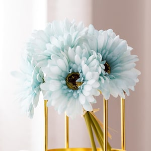 May include: A bunch of light blue gerbera daisies with dark brown centres. The flowers are arranged in a gold-coloured metal vase. The stems are green and tied together with natural-coloured twine.