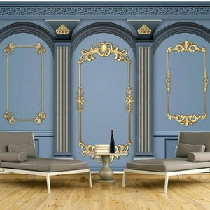 May include: Two gray chaise lounges with white cushions are positioned in front of a blue wall with gold decorative accents. The wall features three ornate gold frames, two of which are empty and one that contains a bottle of wine and a stack of books. The wall is decorated with a decorative molding around the top.