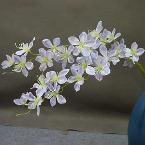 May include: A delicate floral stem featuring multiple pale lavender flowers with green centers and stems. The flowers have translucent petals and are arranged along a curved branch. The background is a muted gray, highlighting the soft colors of the flowers.