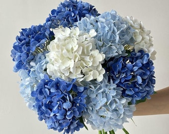 Puede incluir: Un ramo de hortensias azules y blancas. Las flores varían en tonos de azul oscuro a azul claro y blanco. El ramo se mantiene unido por tallos verdes.