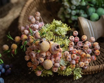 Puede incluir: Primer plano de un arreglo decorativo de bayas y vegetación artificiales. Las bayas son en tonos naranja, rojo y beige, con un aspecto esmerilado. El arreglo incluye pequeñas hojas verdes y está colocado en una cesta tejida.