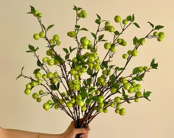 May include: A bouquet of artificial green apple branches with small, round, light green apples and dark green leaves. The branches are held in a hand with a gold bracelet. The background is a solid, light beige color.