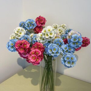 May include: A bouquet of artificial flowers in a clear glass vase. The flowers are a mix of colors, including blue, pink, and white. The flowers have a spherical shape and are clustered together. The vase is sitting on a light yellow surface.