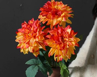 May include: A bouquet of three vibrant, artificial dahlias. The flowers display a gradient of colors, transitioning from a deep red at the tips to a bright yellow at the base. Green leaves and stems complete the arrangement, set against a dark background.