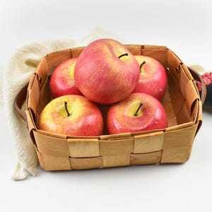 May include: A woven basket containing five red and yellow apples. The apples have a glossy appearance and are arranged in a square basket with a brown handle. The basket is made of light-coloured wood.