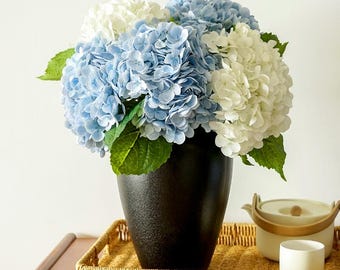 Puede incluir: Un ramo de hortensias azules y blancas con hojas verdes en un jarrón negro. El jarrón está sobre una bandeja tejida, acompañado de una pequeña taza blanca y una tetera blanca con un asa de madera.