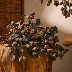 May include: A close-up of a decorative floral arrangement featuring brown, oval-shaped berries and green leaves. The arrangement is placed on a wooden surface, with a natural wood grain texture. The berries are clustered on thin, brown stems.