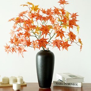May include: A black ceramic vase holds a vibrant arrangement of orange and yellow maple leaves. The vase is cylindrical with a textured surface. Books and small white cups are in the background, creating a decorative display.