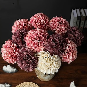 Pode incluir: Um buquê de flores de hortênsia artificiais em tons de rosa, bordô e creme. As flores são dispostas em um vaso rústico e texturizado, criando uma exibição floral vibrante e elegante. O arranjo está sobre uma superfície de madeira escura.